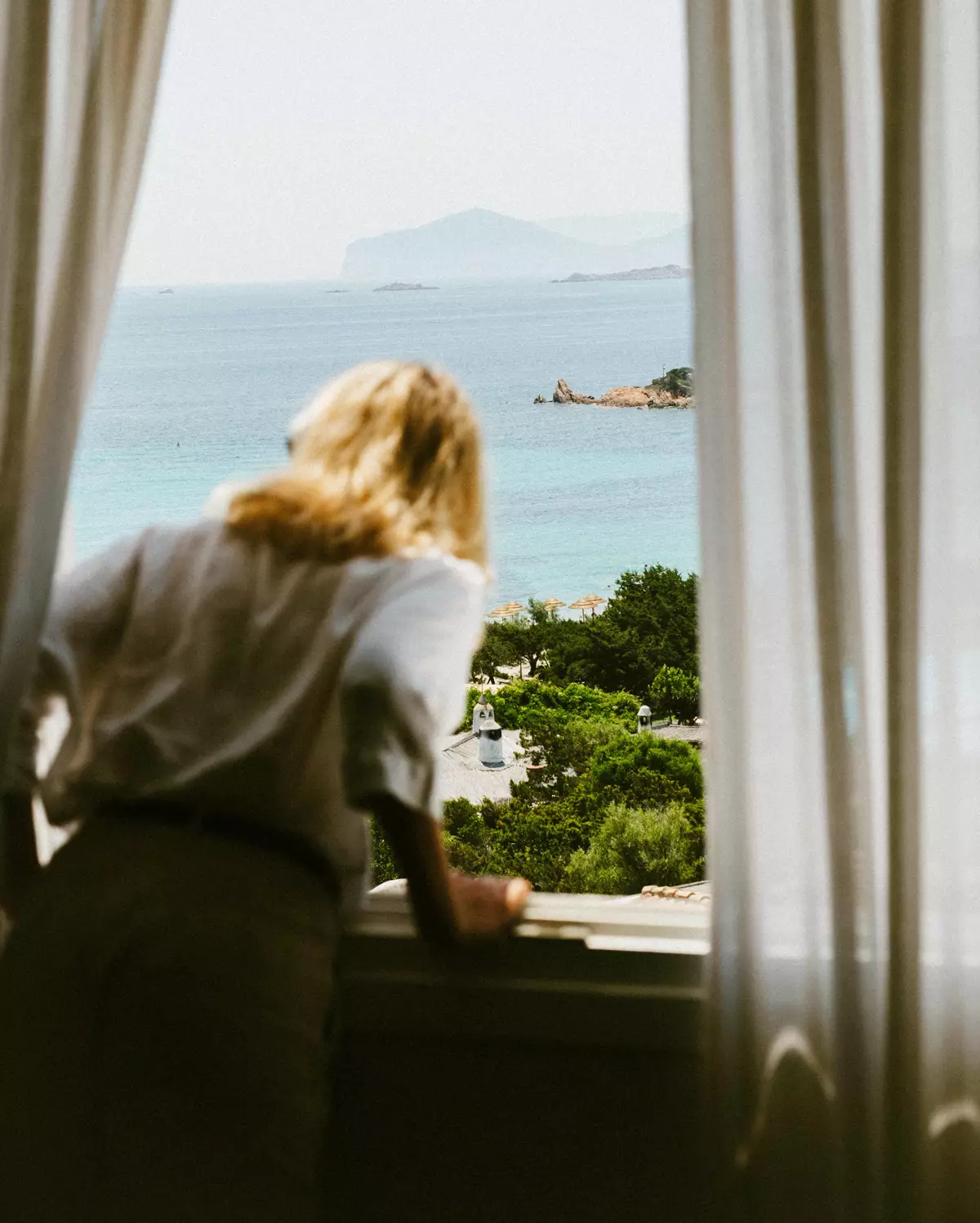 Huésped mirando al mar y a una playa con palapas desde una ventana con cortinas blancas